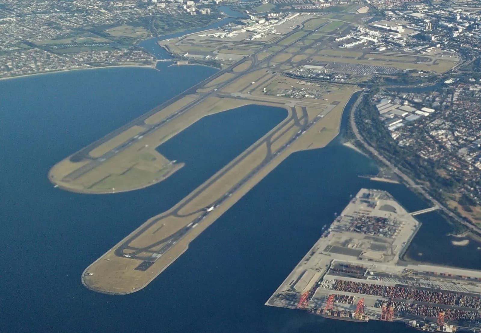 An aerial view shows Sydney Kingsford Smith Airport's parallel runways, terminal buildings, and surrounding city.