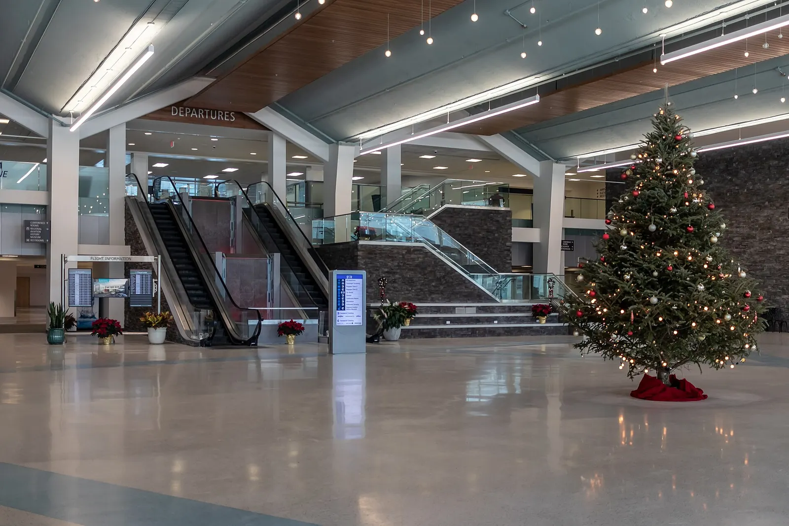 Post-renovation departures hall at Syracuse Hancock International Airport with stone walls, wood ceiling, and the down escalator to bag claim.