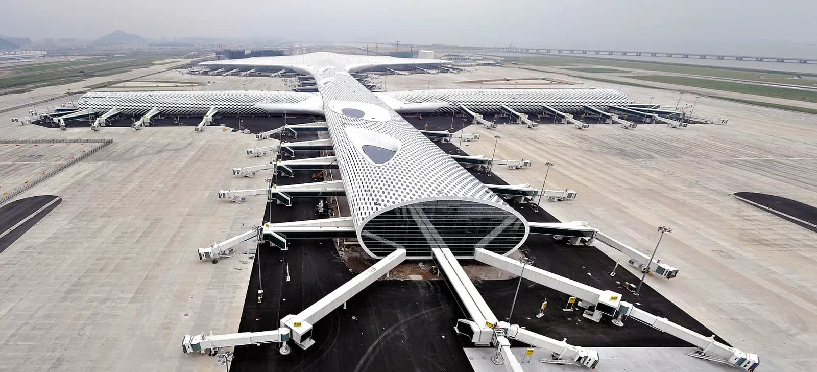 Wide panoramic exterior of Shenzhen Bao'an International Airport Terminal 3 with its distinctive white wave-like roof beside the access road and forecourt.