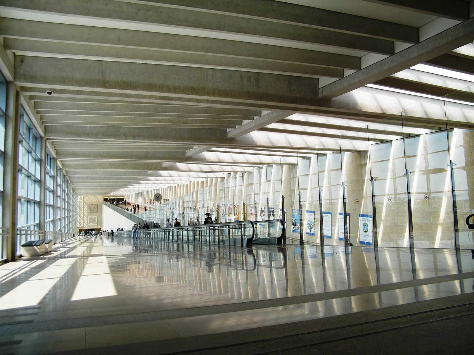 A wide, brightly lit pedestrian walkway inside Ben Gurion International Airport with glass walls and travelers walking toward the gates.