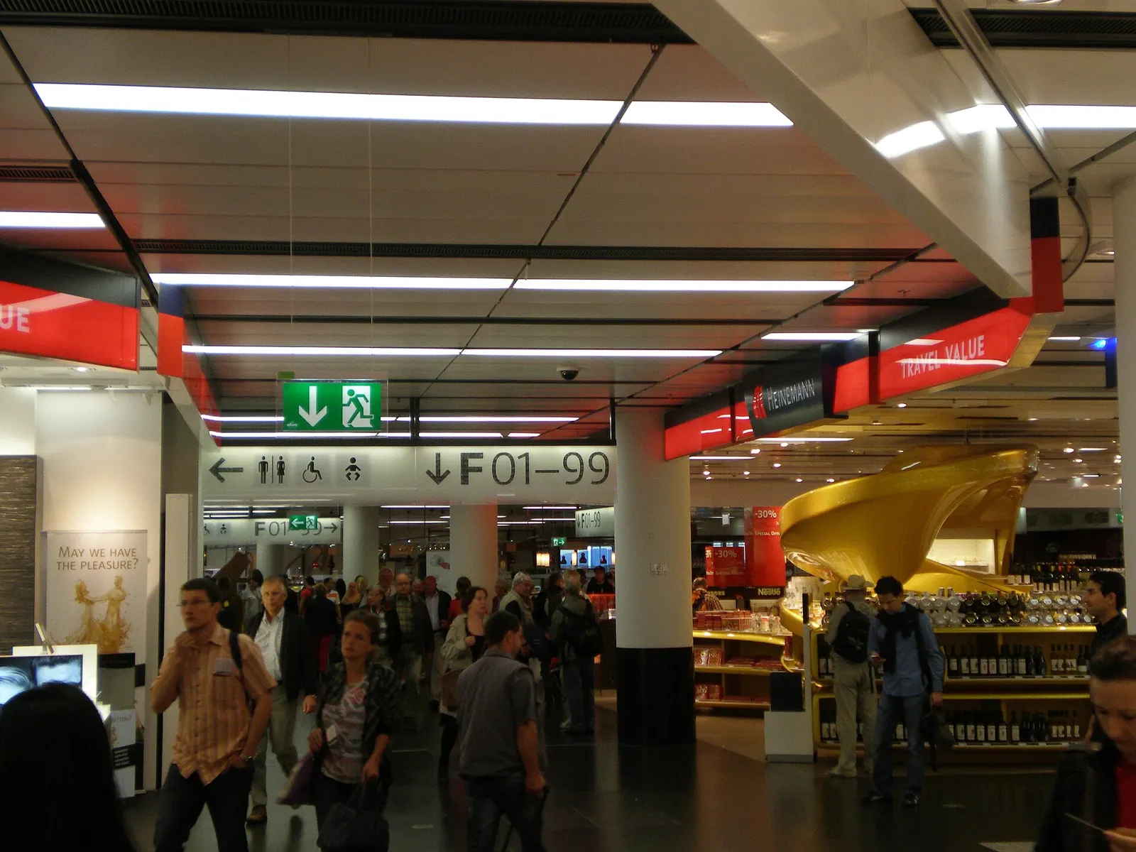 A bright airside duty-free shopping corridor near the F-gates, with retail displays, brand signage, and passengers browsing.