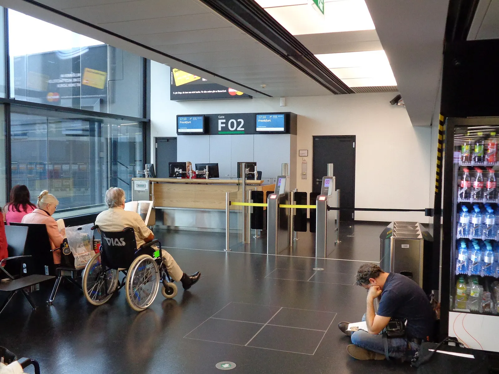 Gate F02 waiting area with rows of seating, large windows overlooking parked aircraft on the tarmac, and a gate number sign.