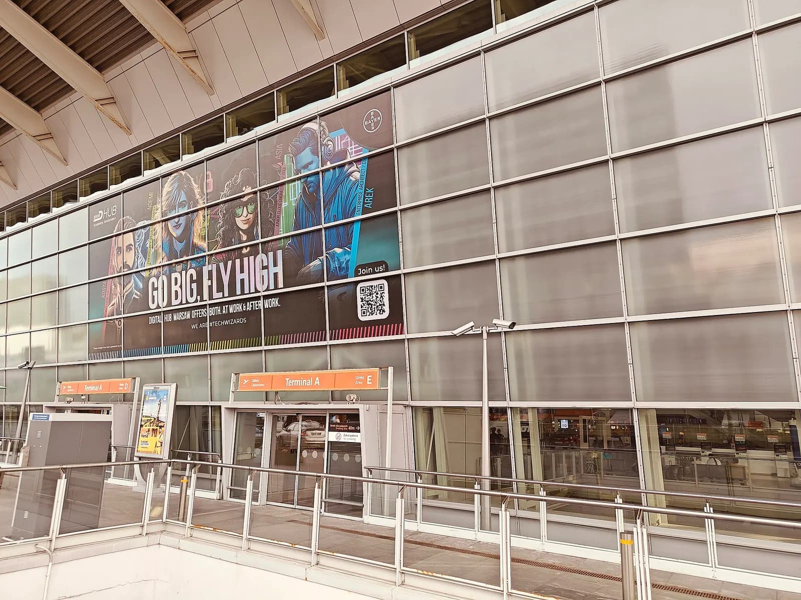 The curved glass-and-steel facade of Terminal A at Warsaw Chopin Airport on a clear summer day, with entrance canopies and landscaping in front.