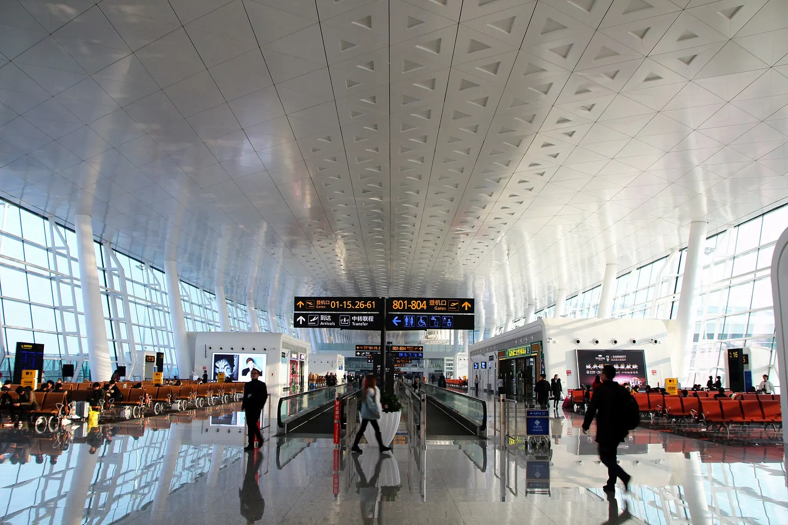 The curved glass-and-steel facade of Wuhan Tianhe International Airport Terminal 3 under a bright sky, with roadways and landscaping in front.