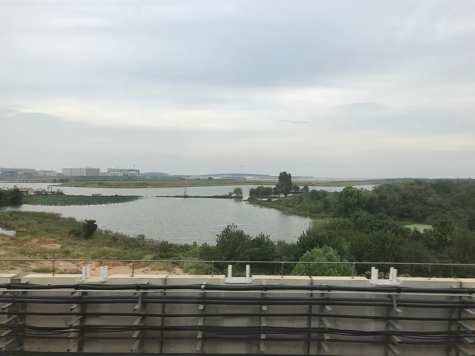 A view from a Wuhan Metro Line 2 train window showing Wuhan Tianhe International Airport buildings and runways beyond a lake.