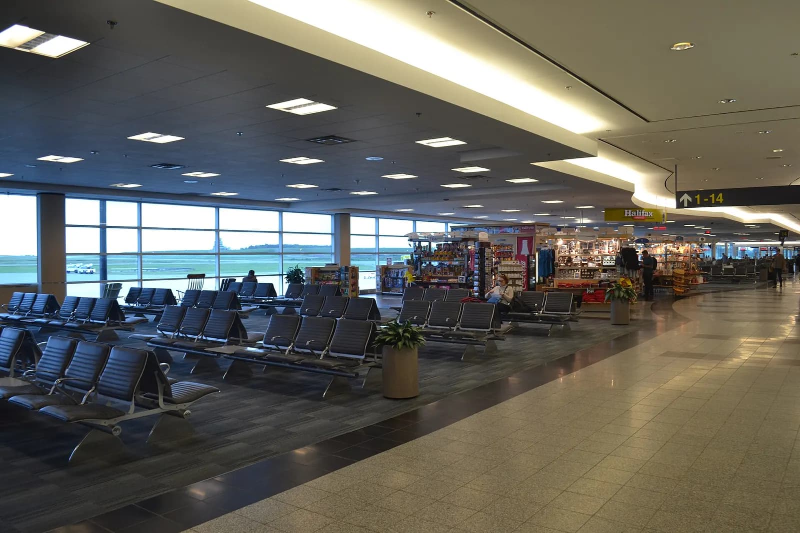 Airside walkway past a Halifax-branded newsstand toward gates 1–14.