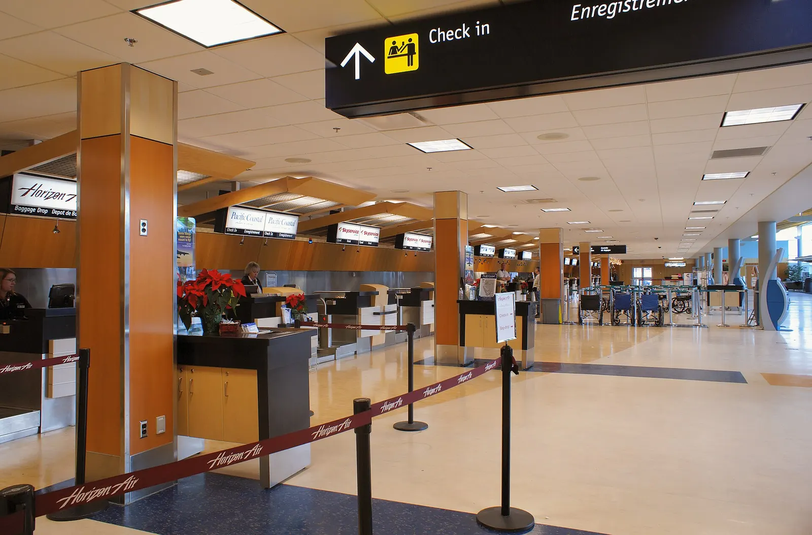 Landside check-in hall at Victoria International Airport with ticket counters, departure boards, and passengers in the queue.