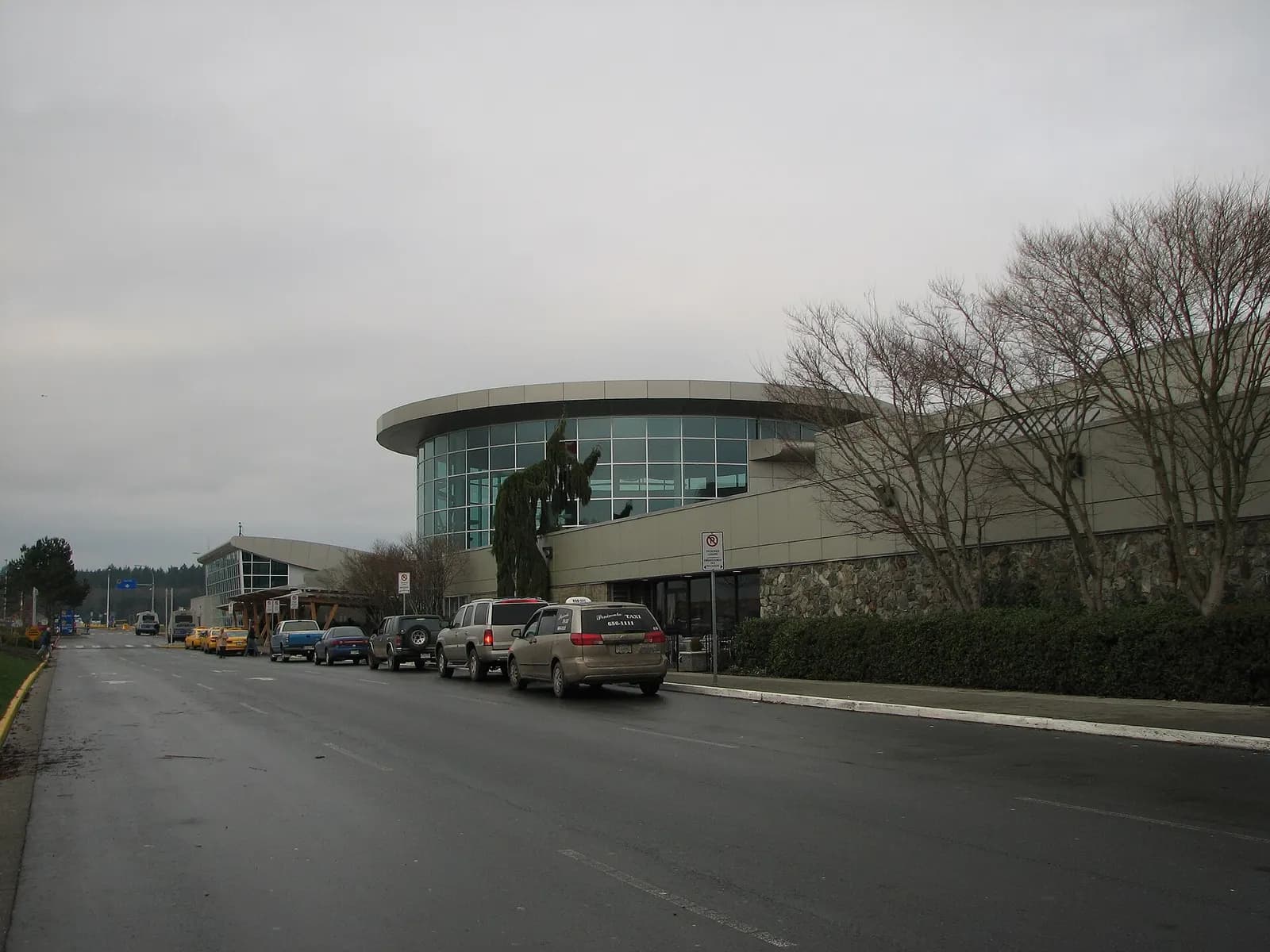 Low brick passenger terminal and control tower at Victoria International Airport under a blue sky, with parked cars in the foreground.