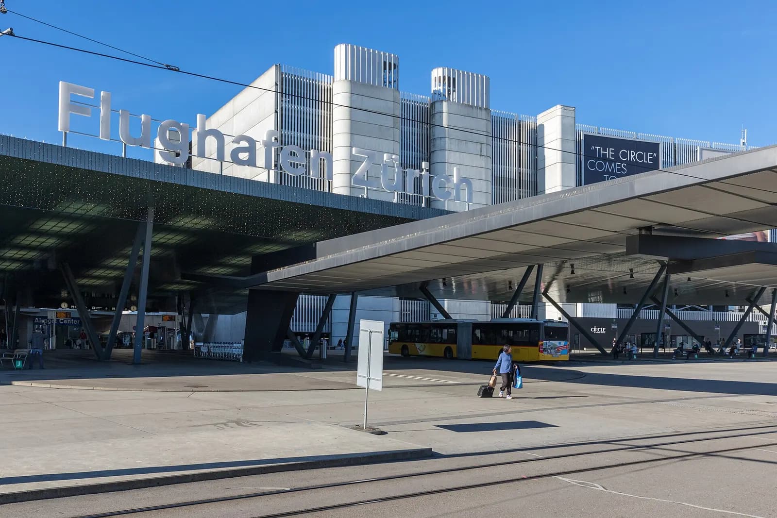 The covered bus and tram stop outside Zurich Airport with multiple bays, signage, and vehicles serving the terminal forecourt.