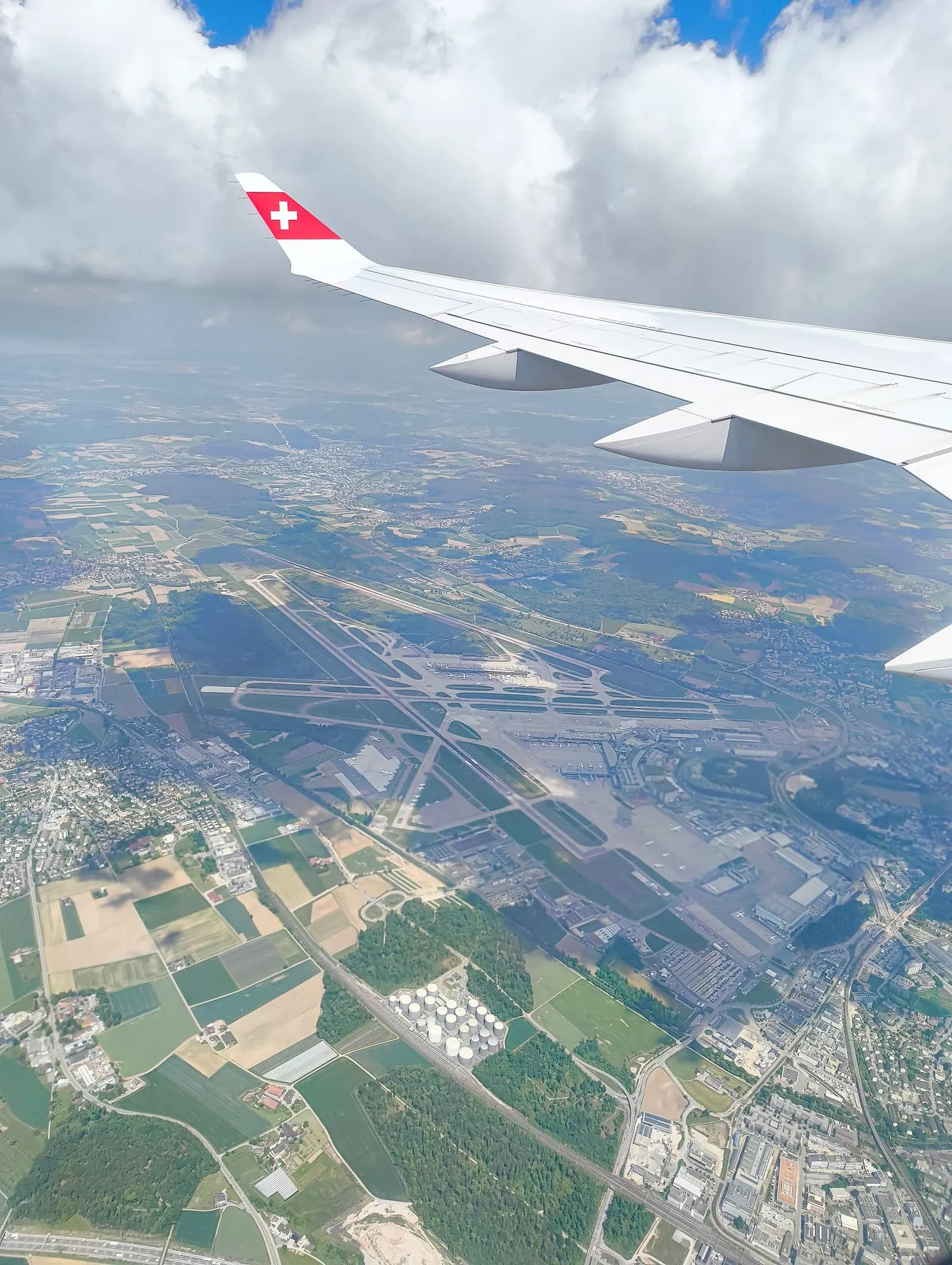 An aerial view of Zurich Airport showing parallel runways, terminal buildings, midfield Dock E, and surrounding green countryside.
