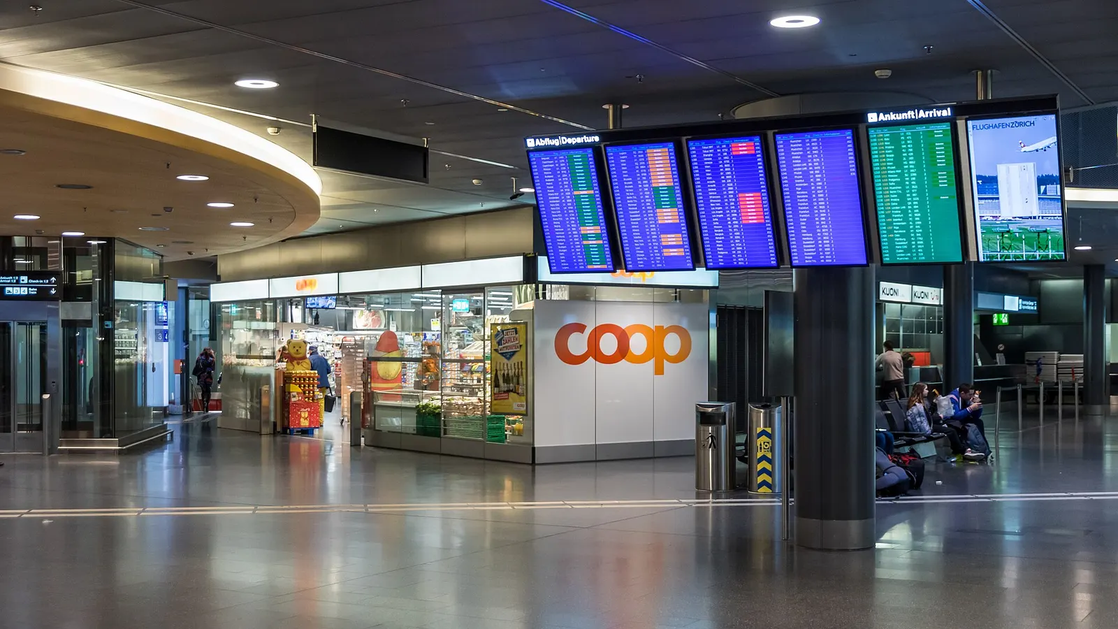 The main check-in hall at Zurich Airport with airline counters, large departure information screens, and a Coop supermarket visible along the concourse.
