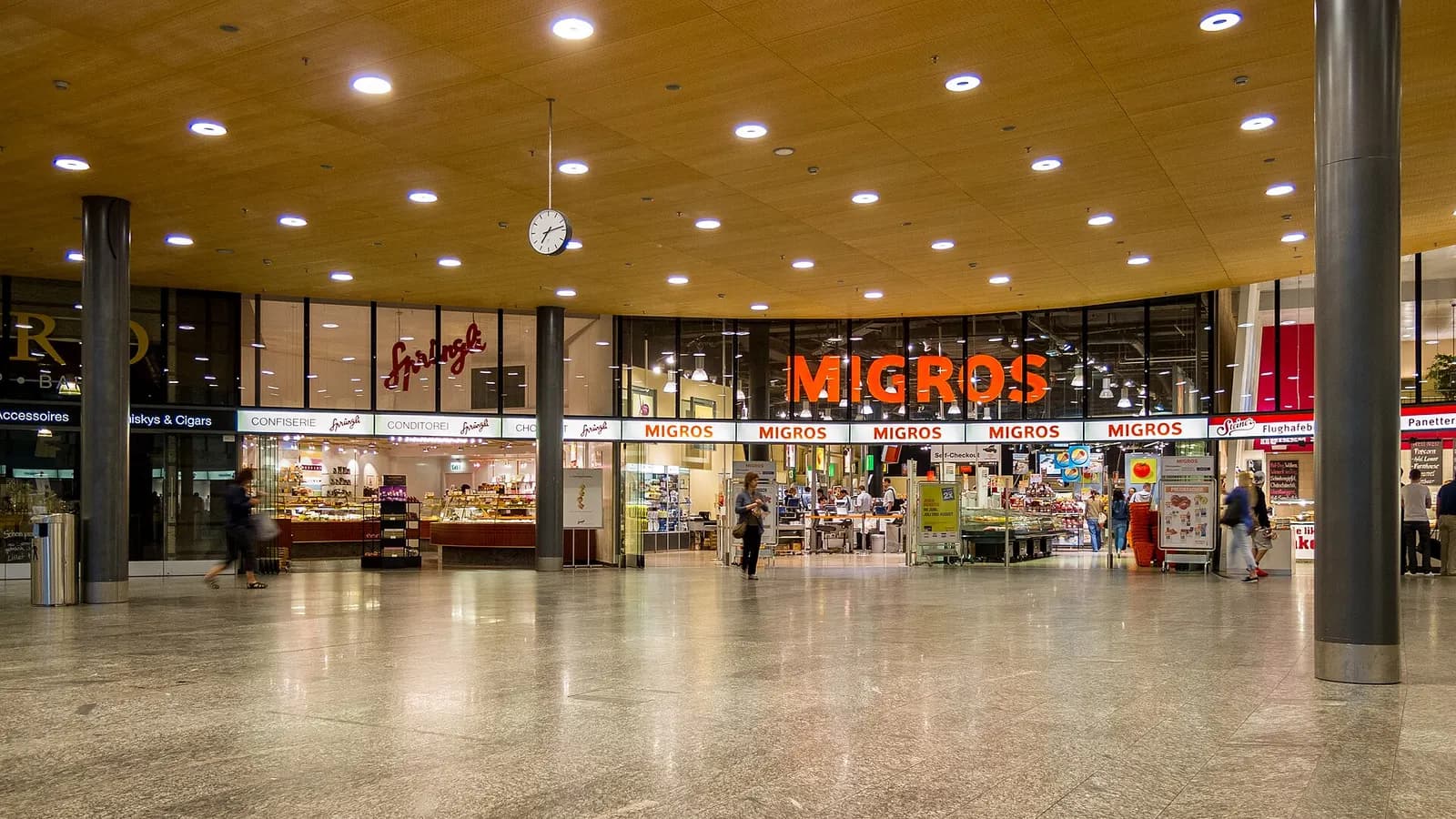 A bright airport shopping corridor with a Migros supermarket, Sprüngli confectionery, and other retail storefronts in the terminal.