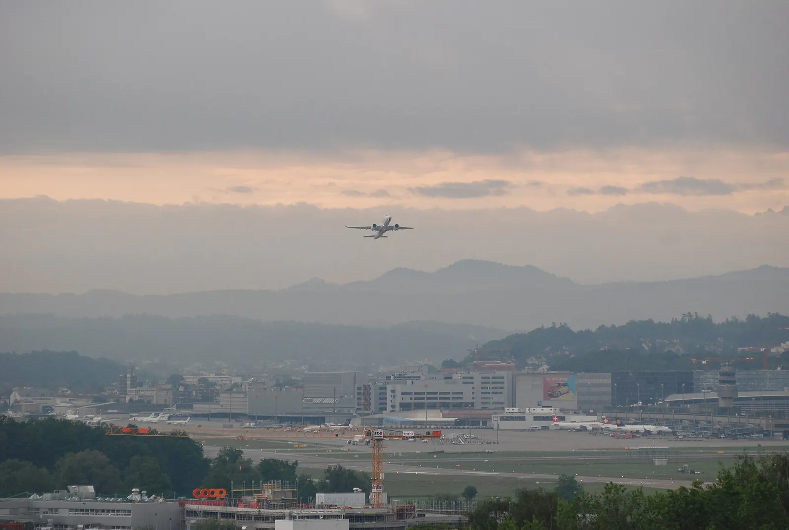 A wide exterior view of Zurich Airport from Rümlang showing runways, cargo facilities, Dock A, the Airside Center, and aircraft taking off.